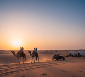A group of friends on a dune during sunset in the Dubai desert with camels and a camp in the background.